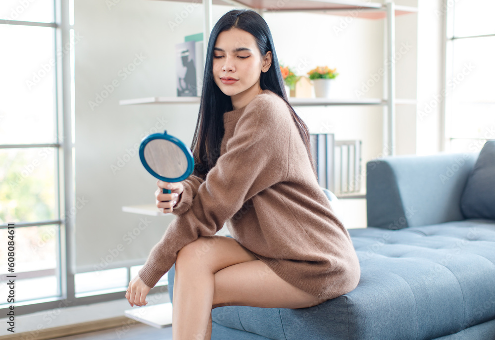 Millennial Asian beautiful pretty female model in casual outfit big brown warm sweater sitting on cozy sofa couch holding handheld mirror posing in living room. Millennial Asian beautiful pretty female model in casual outfit big brown warm sweater sitting on cozy sofa couch holding handheld mirror posing in living room.