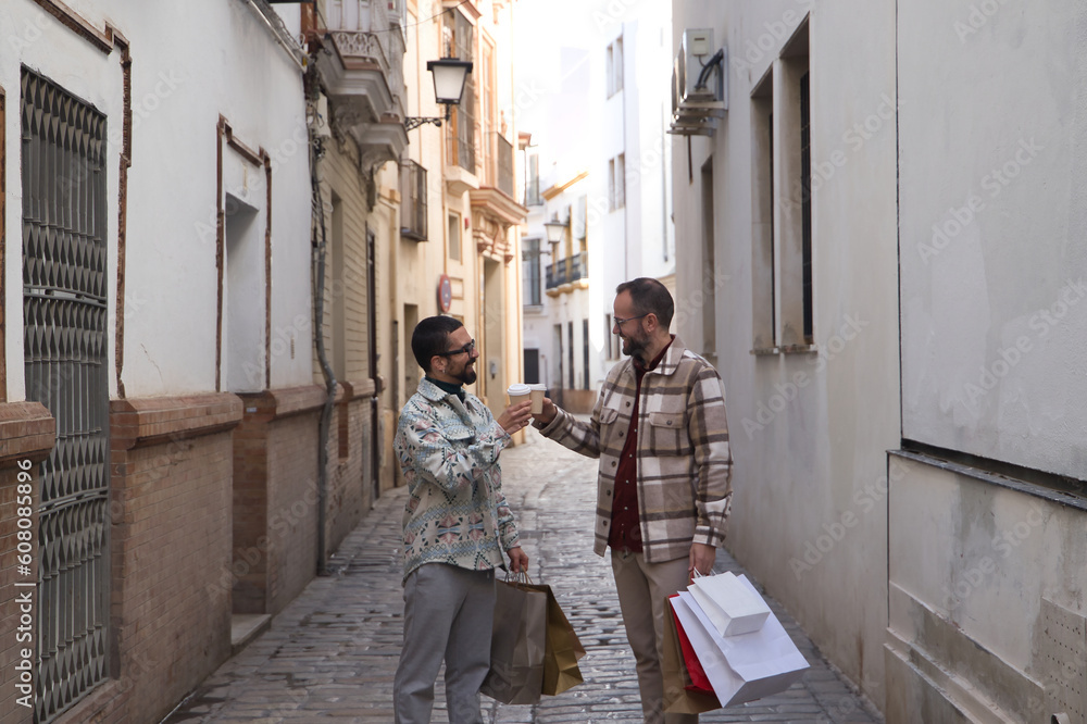Real marriage of gay couple, walking along a lonely street, with a glass of coffee in one hand and the other full of shopping bags. Concept lgtb, lgtbiq+, couples, in love, shopping, sales. Real marriage of gay couple, walking along a lonely street, with a glass of coffee in one hand and the other full of shopping bags. Concept lgtb, lgtbiq+, couples, in love, shopping, sales.