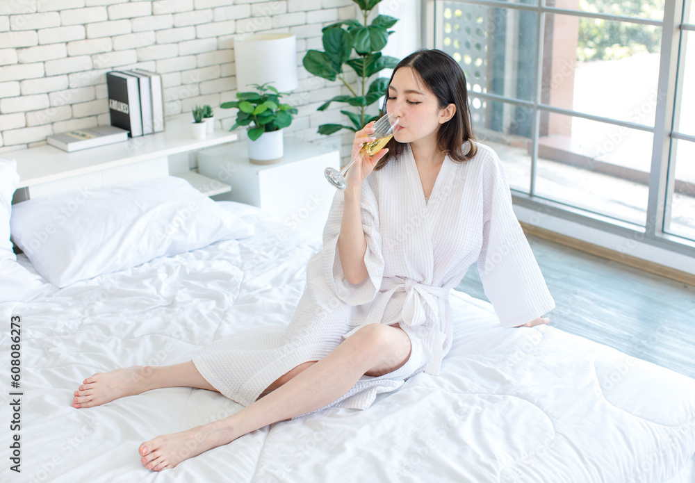 Millennial Asian cheerful happy beautiful female model in casual white clean bathrobe outfit sitting posing smiling holding sparkling champagne tall glass cheer toasting party alone on bed in bedroom Millennial Asian cheerful happy beautiful female model in casual white clean bathrobe outfit sitting posing smiling holding sparkling champagne tall glass cheer toasting party alone on bed in bedroom
