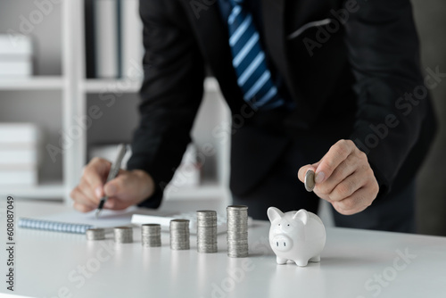 Image of businessman and pile of money showing growth Put the coins in the piggy bank and record the income. expenses on desk money saving concept business finance investment planning.