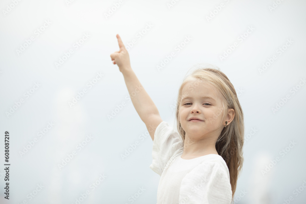 Look there. Smiling little girl on the sky background pointing up with her finger. Focus at her face. Copyspace for your text or logo, banner Look there. Smiling little girl on the sky background pointing up with her finger. Focus at her face. Copyspace for your text or logo, banner