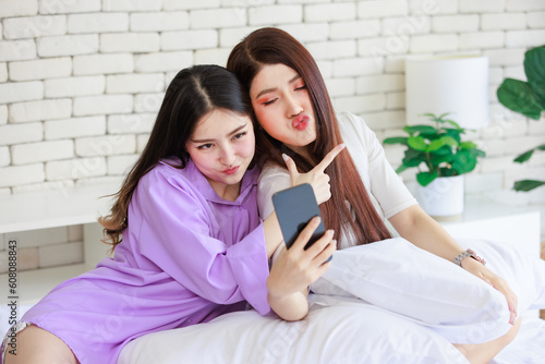 Two Asian cheerful happy female girlfriends in casual outfits sitting resting relaxing holding touchscreen smartphone showing two finger sign smiling taking selfie photo together on bed in bedroom