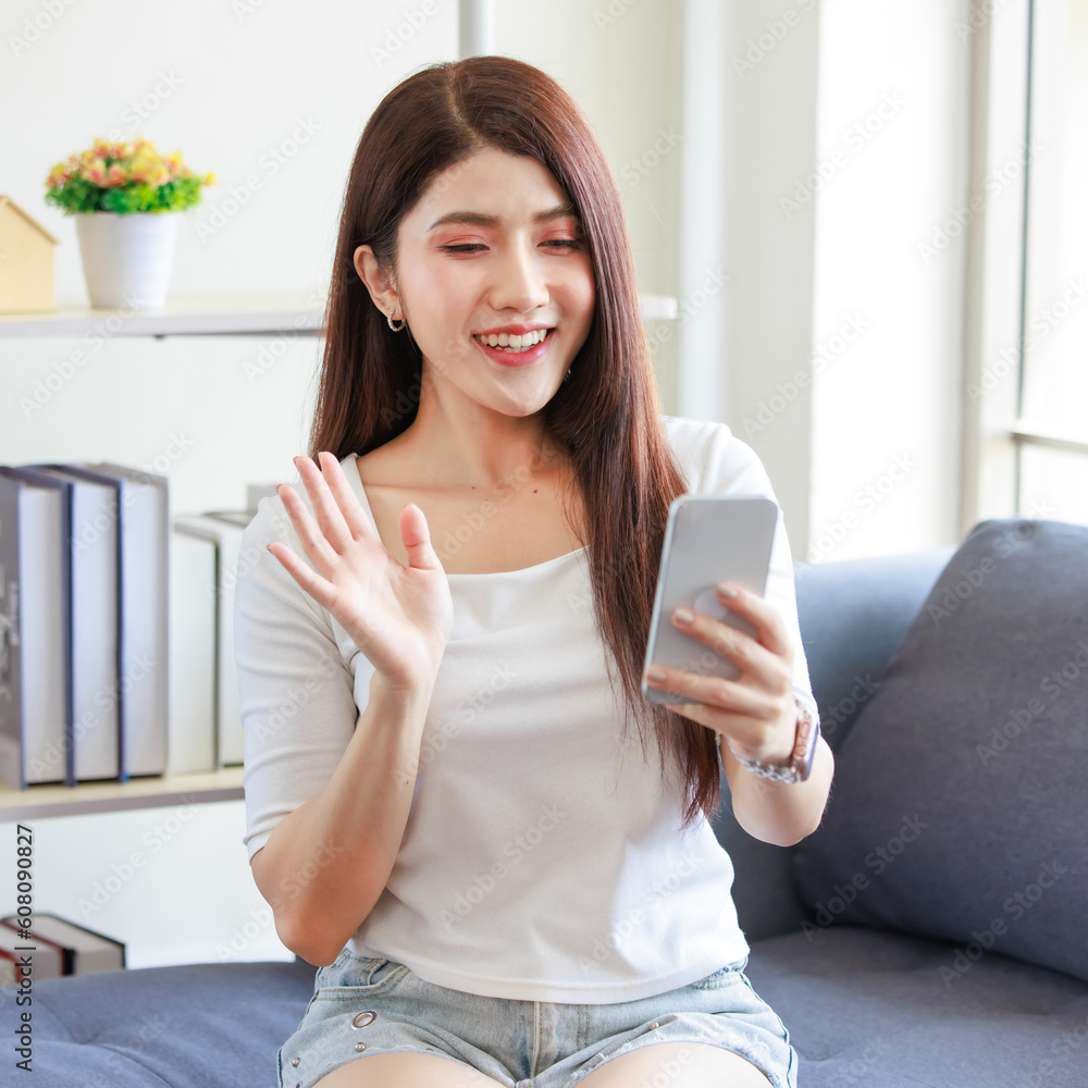 Millennial Asian beautiful lovely female model in casual outfit sitting on cozy sofa couch holding touchscreen smartphone waving hand smiling greeting via online internet video call with friends Millennial Asian beautiful lovely female model in casual outfit sitting on cozy sofa couch holding touchscreen smartphone waving hand smiling greeting via online internet video call with friends