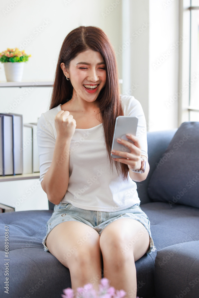 Millennial Asian beautiful lovely female model in casual outfit sitting on cozy sofa couch holding touchscreen smartphone waving hand smiling greeting via online internet video call with friends Millennial Asian beautiful lovely female model in casual outfit sitting on cozy sofa couch holding touchscreen smartphone waving hand smiling greeting via online internet video call with friends