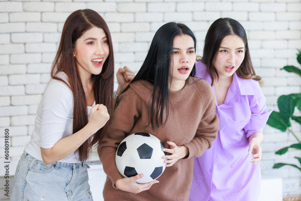 Millennial three Asian beautiful cheerful excited female girlfriends in casual outfit standing smiling holding football exciting focusing while cheering sport soccer team winning cup victory on TV