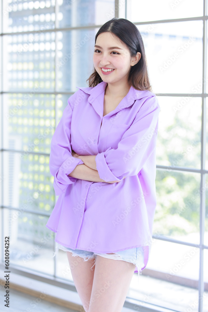 Millennial Asian beautiful cheerful happy female model in casual long sleeve purple shirt outfit and jeans shorts standing crossed arms leaning glass windows smiling to studio camera in living room Millennial Asian beautiful cheerful happy female model in casual long sleeve purple shirt outfit and jeans shorts standing crossed arms leaning glass windows smiling to studio camera in living room
