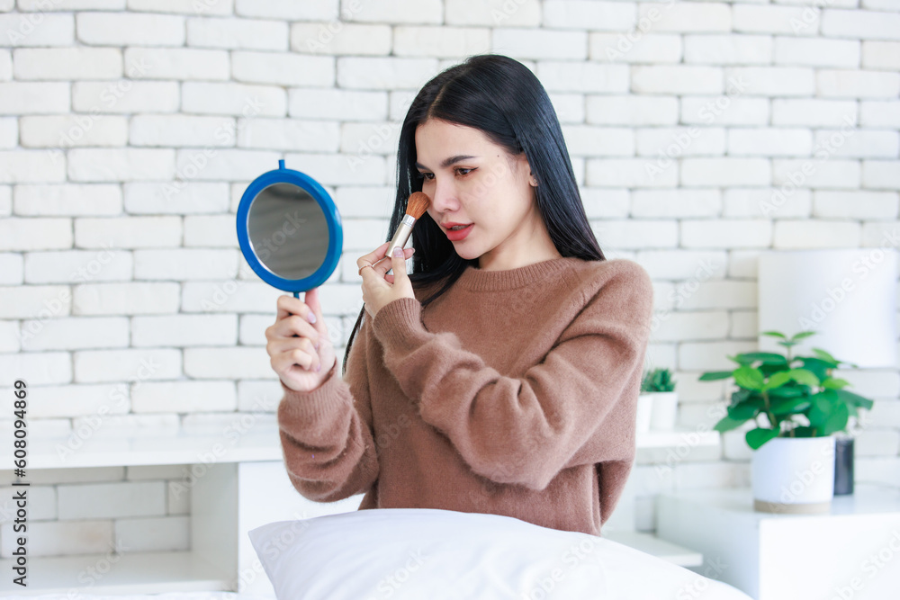 Millennial Asian beautiful cheerful happy female model in casual long sleeve sweater shirt outfit sitting smiling using brush on brushing cosmetic on cheek looking reflection from handheld mirror