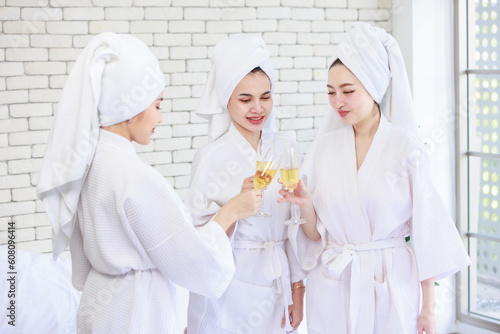 Three Asian cheerful happy beautiful female girlfriends in casual bathrobe and towel outfit standing smiling holding sparkling champagne tall glass cheers toasting together in spa massage studio