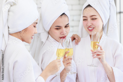 Three Asian cheerful happy beautiful female girlfriends in casual bathrobe and towel outfit standing smiling holding sparkling champagne tall glass cheers toasting together in spa massage studio