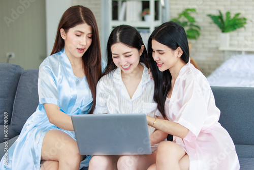 Three Asian cheerful happy female girlfriends in casual pajamas outfit sitting smiling watching surfing browsing internet online via laptop computer party together while drinking sparkling champagne