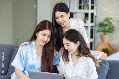Three Asian cheerful happy female girlfriends in casual pajamas outfit sitting smiling watching surfing browsing internet online via laptop computer party together while drinking sparkling champagne