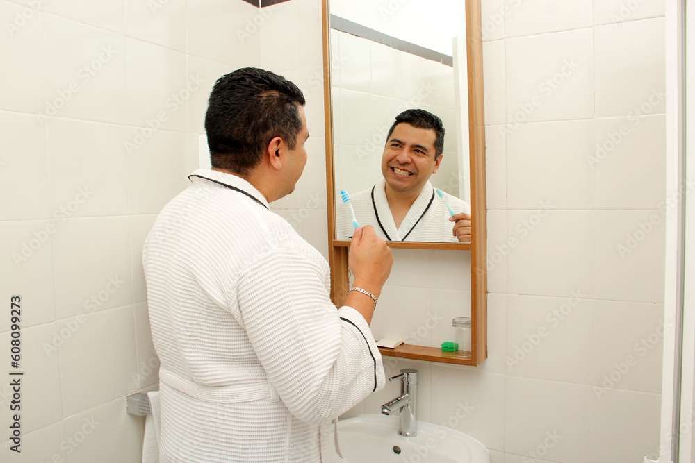 Fototapeta premium Forty-year-old dark-skinned Latino man brushes his teeth in the bathroom in a bathrobe looks in the mirror and takes care of his oral health