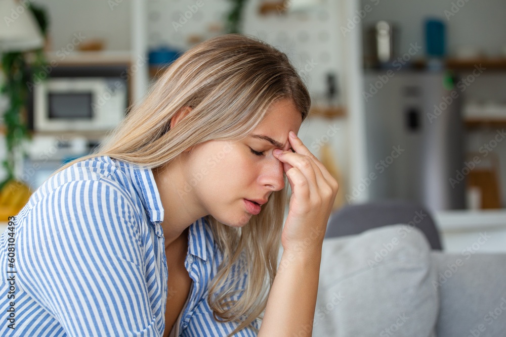 Portrait of a young woman sitting on the couch at home with a headache and pain. Beautiful woman suffering from chronic daily headaches. Sad woman holding her head because sinus pain Portrait of a young woman sitting on the couch at home with a headache and pain. Beautiful woman suffering from chronic daily headaches. Sad woman holding her head because sinus pain