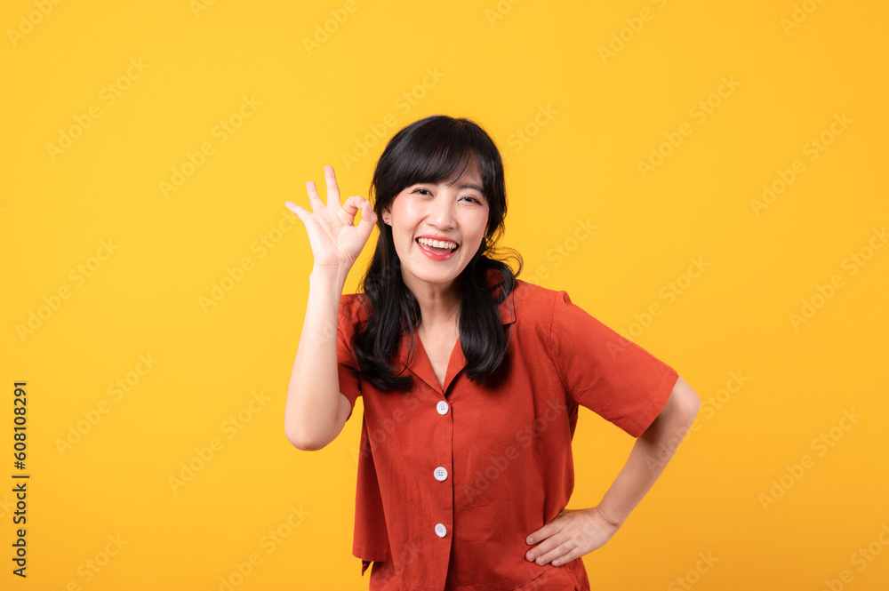 Portrait young beautiful asian woman happy smile dressed in orange clothes looking surprised, reacting amazed, raising eyebrows impressed isolated on yellow studio background.