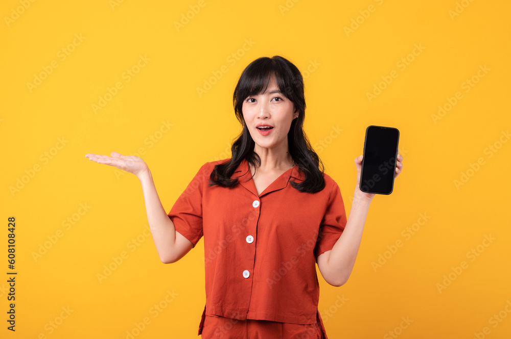 Portrait beautiful young asian woman happy smile dressed in orange clothes with smartphone and pointing hand gesture to free space isolated on yellow studio background. app smartphone concept Portrait beautiful young asian woman happy smile dressed in orange clothes with smartphone and pointing hand gesture to free space isolated on yellow studio background. app smartphone concept