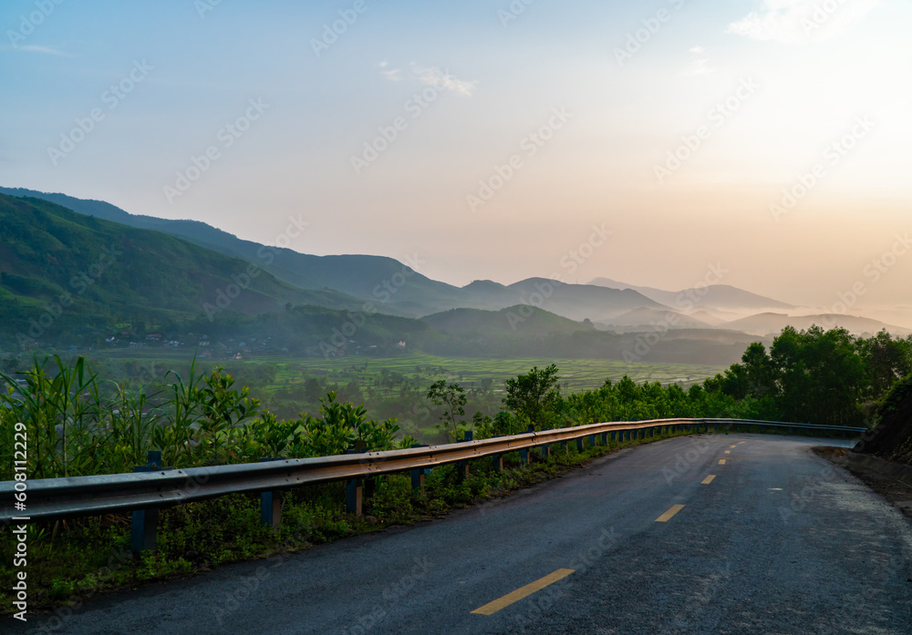 Landscape photo of mountain pass road with trees and valleys, village fields below at dawn.