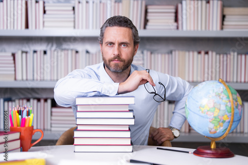 Portrait of school teacher with book in classroom. Handsome teacher in classroom. Teachers Day. Good school teacher. Tutor at classroom. Man with books in classroom. Knowledge and education concept.