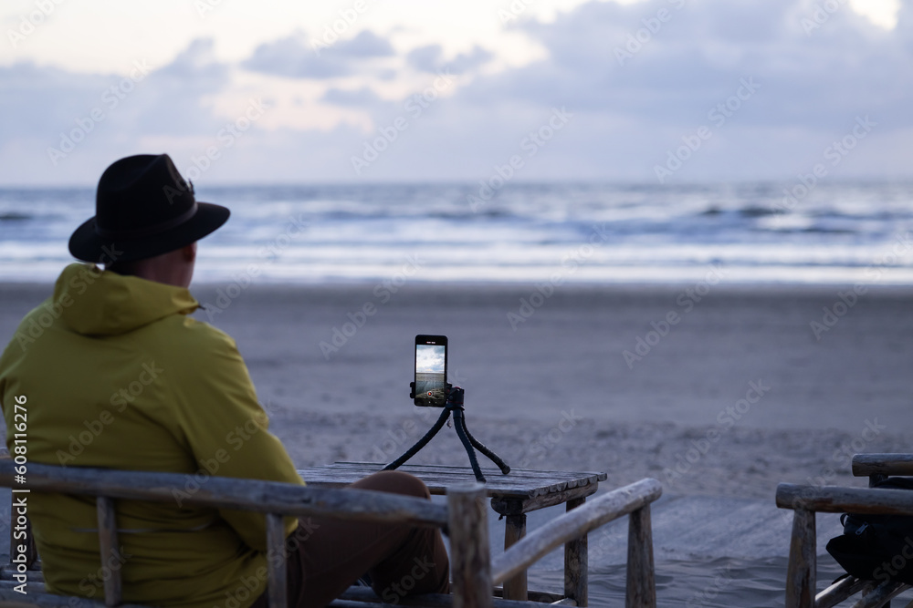 Man in yellow jacket in beach cafe sits on wooden chair, shoots sunset at sea on smartphone