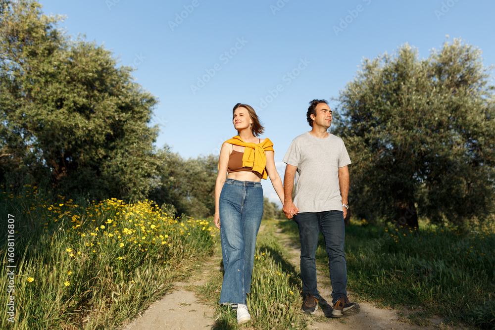 Fototapeta premium Outdoor shot of young couple in love walking on pathway through grass field. Man and woman walking along tall grass field and olive trees