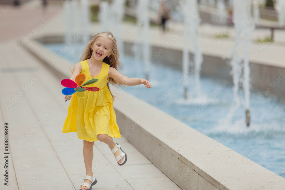 Happy little girl running in the park near the city fountain playing with paper windmill. Summer concept. happy childhood Happy little girl running in the park near the city fountain playing with paper windmill. Summer concept. happy childhood