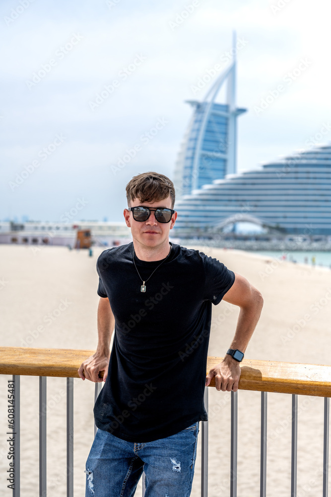 Portrait of young man in casual summer wear with sunglasses leaning on fence and posing on the beach in Dubai.
