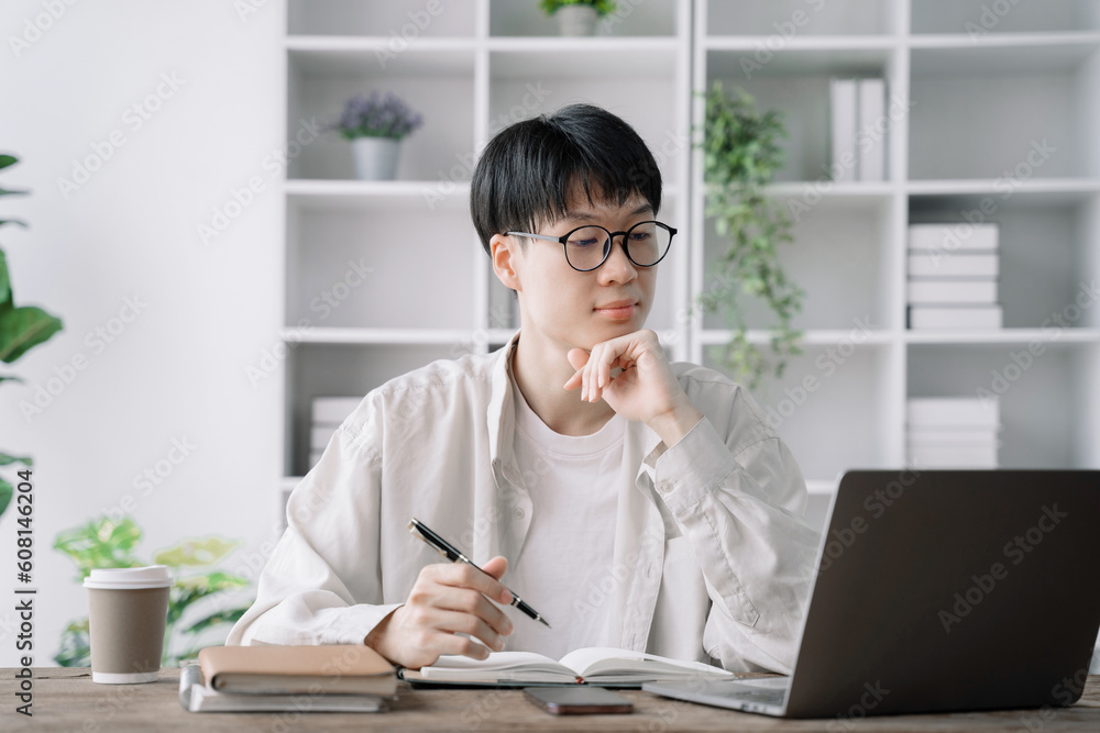 male student in casual clothes sitting at wooden table with laptop and writing notes while preparing for exam in library.