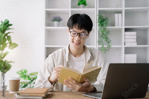 male student in casual clothes sitting at wooden table with laptop and writing notes while preparing for exam in library.