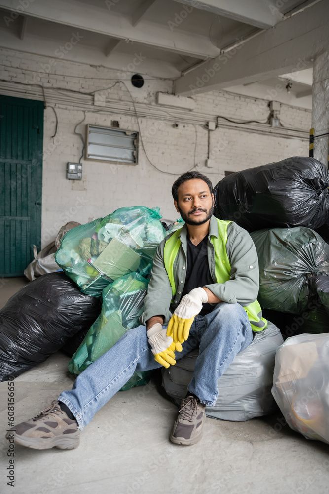 Male indian worker in high visibility vest and protective gloves sitting on plastic bags with trash while working in garbage sorting center, recycling concept Male indian worker in high visibility vest and protective gloves sitting on plastic bags with trash while working in garbage sorting center, recycling concept
