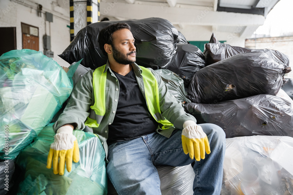 Bearded indian sorter in fluorescent vest and protective gloves looking away while sitting on plastic bags with trash in garbage sorting center, recycling concept