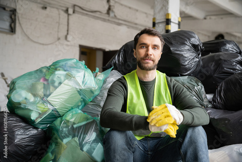 Bearded sorter in protective vest and gloves looking at camera while sitting near plastic bags with trash while working in blurred garbage sorting center, recycling concept