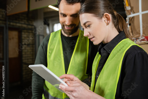 Young worker in safety reflective vest using digital tablet while standing near smiling colleague and blurred waste paper in garbage sorting center, waste sorting and recycling concept