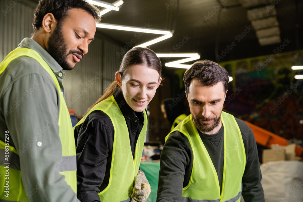Cheerful workers in high visibility vests looking away while indian colleague talking and standing together in blurred garbage sorting center, garbage sorting and recycling concept Cheerful workers in high visibility vests looking away while indian colleague talking and standing together in blurred garbage sorting center, garbage sorting and recycling concept