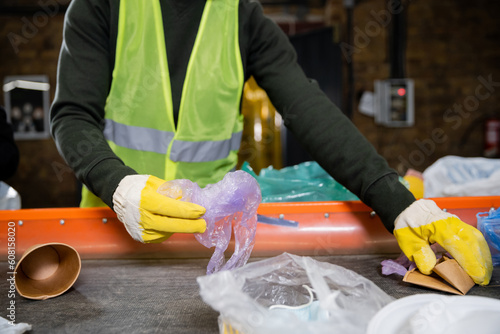 Cropped view of sorter in protective gloves and vest taking garbage from conveyor while working in blurred garbage sorting center, garbage sorting and recycling concept