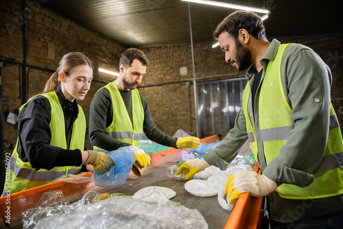 Multiethnic workers in protective vests and gloves taking trash from conveyor while working together in waste disposal station at background, garbage sorting and recycling concept