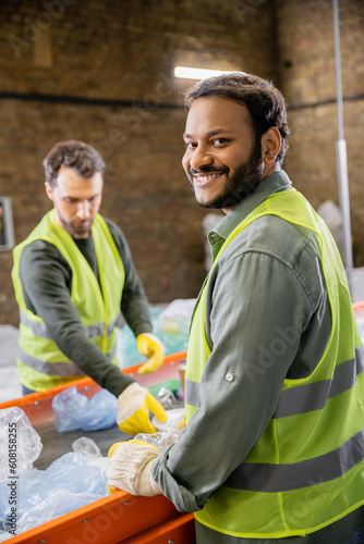 Positive indian worker in protective clothes looking at camera while standing near trash on conveyor and blurred colleague in garbage sorting center, garbage sorting and recycling concept © LIGHTFIELD STUDIOS