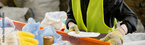 Cropped view of sorter in protective vest and gloves taking garbage from conveyor while working in waste disposal station at background, garbage sorting and recycling concept, banner