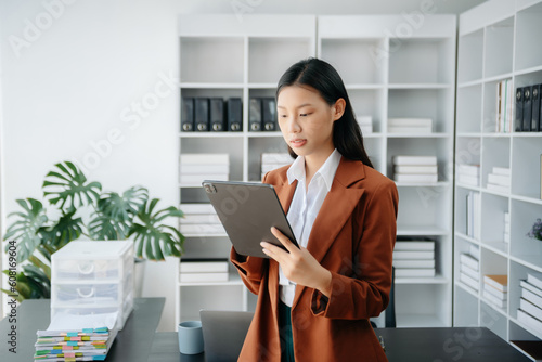  Beautiful Asian woman using laptop and tablet while sitting at her working place. Concentrated at work.