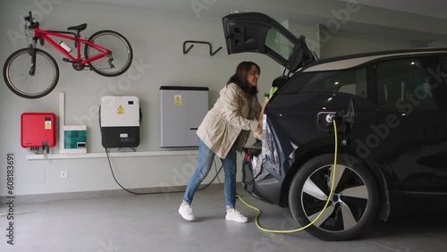 Woman parks in the garage and plugs a charger into socket of the electric car. Renewable houses, Energy Efficient Domestic Life. Horizontal Video 4k.