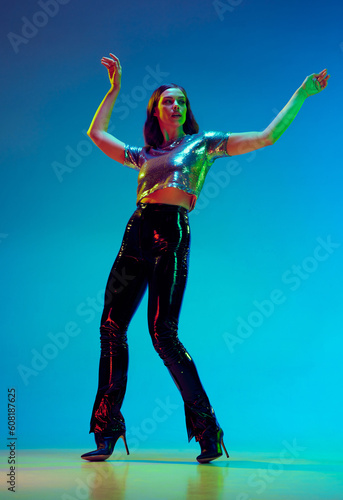 One woman, young fashion model wearing fashionable clothes with leather black pants and silver top posing with over blue background in neon light. Concept of beauty, fashion week