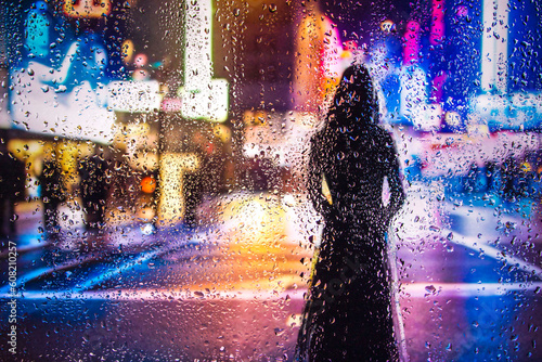 iew through glass window with rain drops on blurred reflection silhouette of a girl on a city street after rain and colorful neon bokeh city lights, night street scene. Focus on raindrops on glass	 photo
