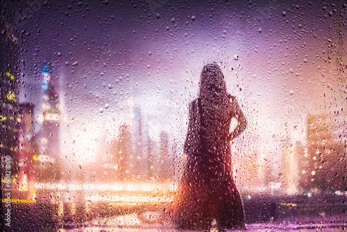 Fototapeta Naklejka Na Ścianę i Meble -  View through glass window with rain drops on blurred reflection silhouette of a girl on a city street after rain and colorful neon bokeh city lights, night street scene. Focus on raindrops on glass