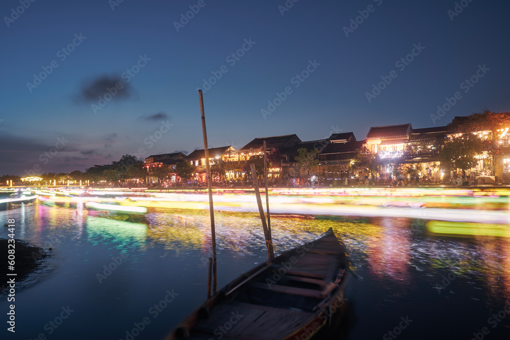 Fototapeta premium Light trails of boats with traditional lanterns on the motion. Night scene of water canal and waterfront in ancient city Hoi An in Vietnam..