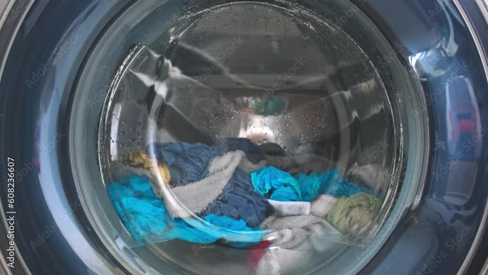 The washing machine washes dirty colored clothes. Washing clothes in a household washing machine. Close-up of the rotation of a drum washing machine. The washing machine washes dirty colored clothes. Washing clothes in a household washing machine. Close-up of the rotation of a drum washing machine.