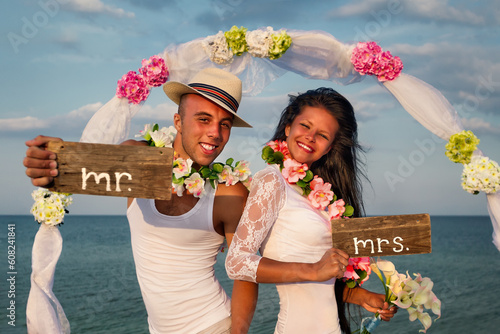 Groom with bride wearing lei, standing under archway on beach photo
