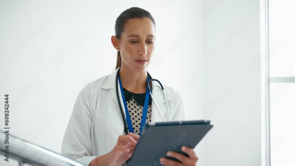 Portrait of female doctor wearing white coat walking into focus looking at patient notes on digital tablet on hospital stairs - shot in slow motion Portrait of female doctor wearing white coat walking into focus looking at patient notes on digital tablet on hospital stairs - shot in slow motion