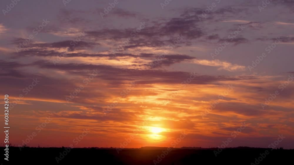 Fiery Yellow Sun Sets over Horizon in a Field in Amazing Dramatic Orange Blue Lilac Sky with Timelapse Clouds. Beautiful Landscape of Sunset, Setting Sun in a Field with Colorful Clouds in Sunlight.
