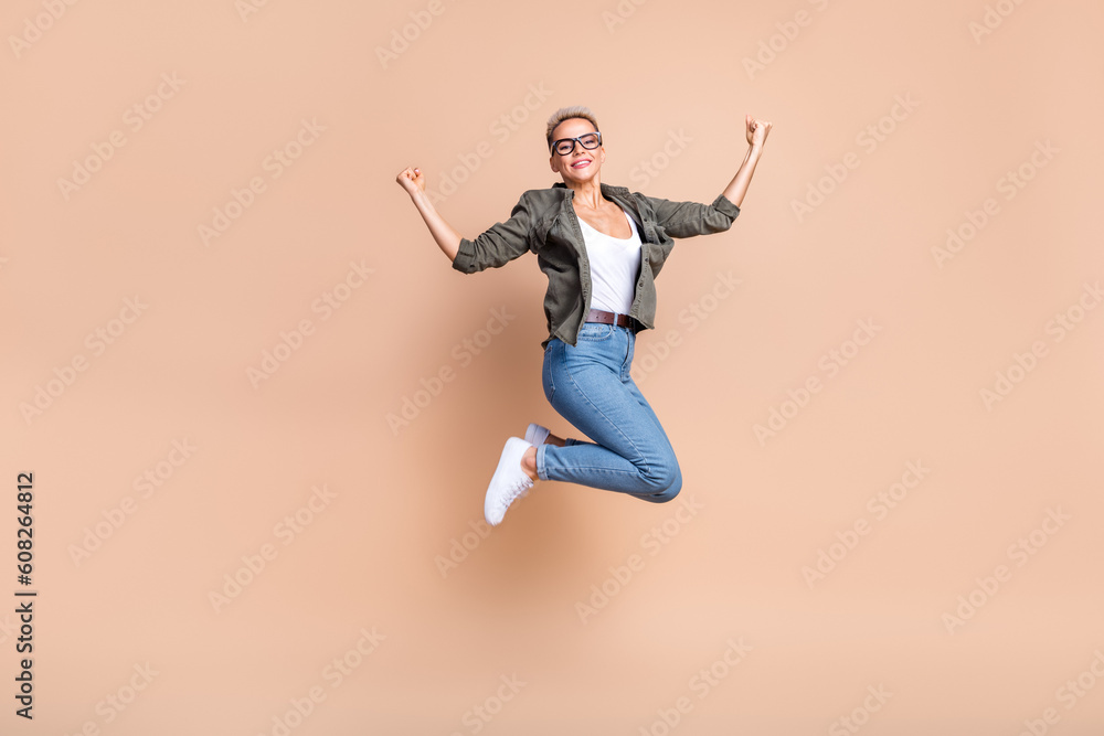 Full length photo of excited lucky lady dressed khaki shirt jumping high rising fists isolated beige color background Full length photo of excited lucky lady dressed khaki shirt jumping high rising fists isolated beige color background