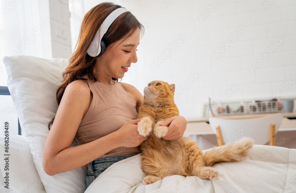 Pretty Asian woman with headphone hold and hug orange cat sit on the bed in bedroom with smiling and cat look at woman face. Pretty Asian woman with headphone hold and hug orange cat sit on the bed in bedroom with smiling and cat look at woman face.