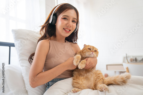 Lovely Asian girl with headphone hold and cudding orange cat and sit on bed also look at camera with smiling and day light.