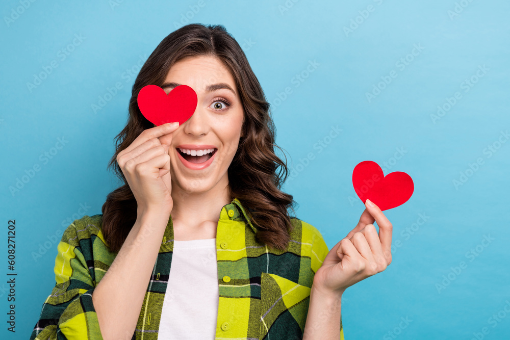 Portrait of positive funky lady arms hold small paper heart cover one eye isolated on blue color background Portrait of positive funky lady arms hold small paper heart cover one eye isolated on blue color background